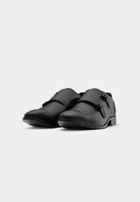 Et par sorte læder monk strap dresssko med dobbelte spænder på en hvid baggrund, set fra en vinkel.