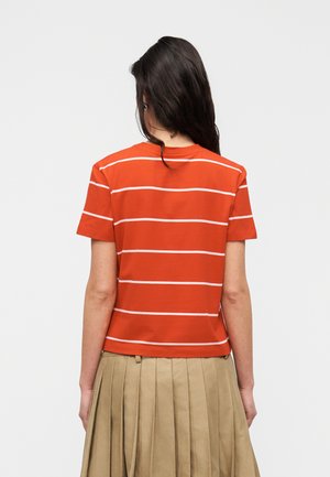 Femme aux longs cheveux foncés portant un t-shirt rouge à manches courtes avec des rayures blanches horizontales et une jupe plissée beige, vue de dos.