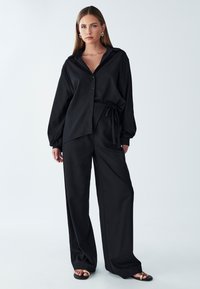 Camicia nera a maniche lunghe con bottoni e cintura in vita, abbinata a pantaloni neri larghi. Tessuto morbido, design minimalista e senza fantasie.