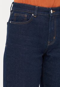 Džínové kalhoty v tmavě modré barvě s žlutým prošíváním, střih s těsným designem, tradiční pětipocket styl a kovový knoflík na zapínání v pase.