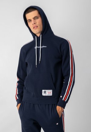 Sudadera color azul marino de algodón, con franjas laterales en blanco y rojo, bolsillo delantero tipo canguro y un logo de Champion en el pecho.