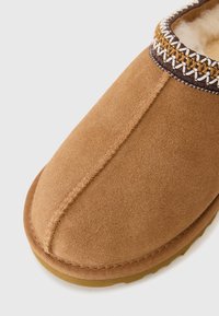 Pantofole in suede marrone con punta arrotondata, caratterizzate da una rifinitura decorativa cucita in marrone e oro, e una morbida fodera in pelliccia.