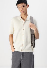 Polo en maille beige clair à manches courtes, avec un motif texturé et des boutons marron, associé à un pantalon large gris foncé.