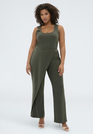 Donna con capelli ricci che indossa una tuta verde oliva senza maniche con un dettaglio gonna incrociata asimmetrica e tacchi beige open toe.