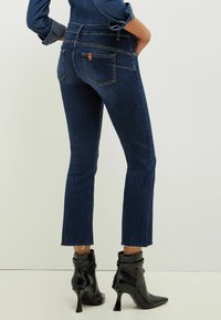 Tmavě modré zvonové džíny s netříděným lemem, se zadními kapsami a jemným prošitím. Kombinované s černými vysokými kotníkovými botami.