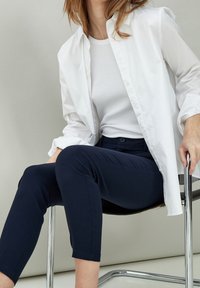 Chemise blanche boutonnée portée sur un débardeur blanc, associée à un pantalon ajusté bleu marine. Mannequin assis sur une chaise chromée sur fond clair.