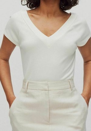 Persoon draagt een wit V-hals top met korte mouwen, ingestopt in een beige broek met hoge taille, handen in de zakken, tegen een effen achtergrond.