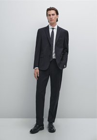 Sort jakkesæt med skræddersyet pasform, der består af en enkeltbrystet jakke med revers og matchende bukser. Kombineret med en hvid skjorte og sorte sko.