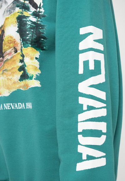 Felpa color teal con una stampa di paesaggio ad acquerello sul davanti. La manica destra presenta un testo bianco audace che dice "NEVADA."