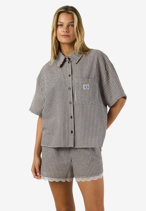 Ženska, oblečena v rjavo-belo srajco s kratkimi rokavi in karirastim vzorcem ter sprednjim žepom, ki se ujema s kratkimi hlačami z čipkastim robom.