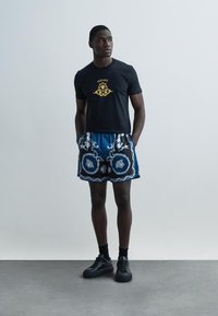 Camiseta preta com logótipo dourado, combinada com calções azuis estampados com desenhos intrincados. Tênis pretos completam o visual.