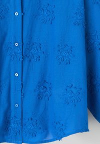 Blouse bleue brodée avec des motifs floraux et des détails de fils texturés. Des boutons descendent sur le devant, mettant en valeur le design.