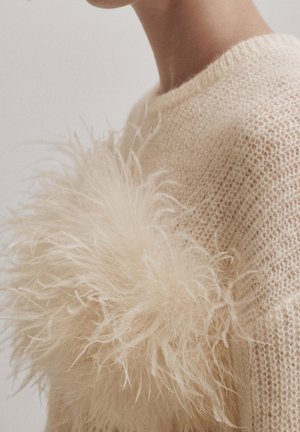 Nahaufnahme einer Person, die einen beigen Strickpullover mit großen, weichen, flauschigen Federverzierungen am Oberarm und Schulterbereich trägt.