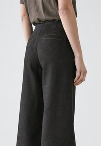 Pantalon à jambes larges, gris foncé, en tissu texturé, avec deux poches arrière et une ceinture nette, présentant une silhouette droite.