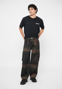 Jovem com cabelo encaracolado a usar uma t-shirt preta Double V, calças cargo em estampa tie-dye castanho e preto, cinto preto e sapatilhas pretas, de pé.