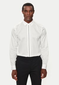 Camicia bianca a maniche lunghe con patta frontale con piping nero, colletto classico e polsini a bottoni, indossata con pantaloni neri.