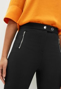 Pantalons noirs ajustés avec poches zippées métalliques et fermeture à pression. Le haut est orange avec une texture douce et des manches retroussées.