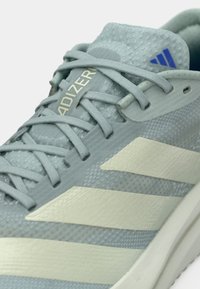 Κοντινή λήψη αθλητικού παπουτσιού Adidas Adizero σε ανοιχτό γκρι, με δεμένα κορδόνια, υφή στην επιφάνεια και κρεμ-χρώματος ρίγες στο πλάι.