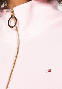 Sudadera rosa con cremallera fabricada en tejido suave. Presenta una cremallera dorada con un tirador circular y un pequeño logo en el pecho.