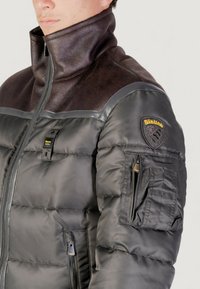 Uomo che indossa una giacca nera imbottita con colletto in pelle e una toppa sulla manica con l'etichetta "Blauer", con tasche con zip visibili sul braccio e sul davanti.