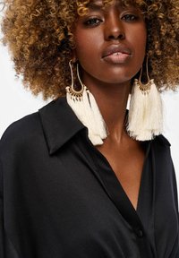 Gouden hoops met witte kwastjes, een zwarte satijnen blouse met een kraag en knoopsluiting, zachte textuur en strakke ontwerp.