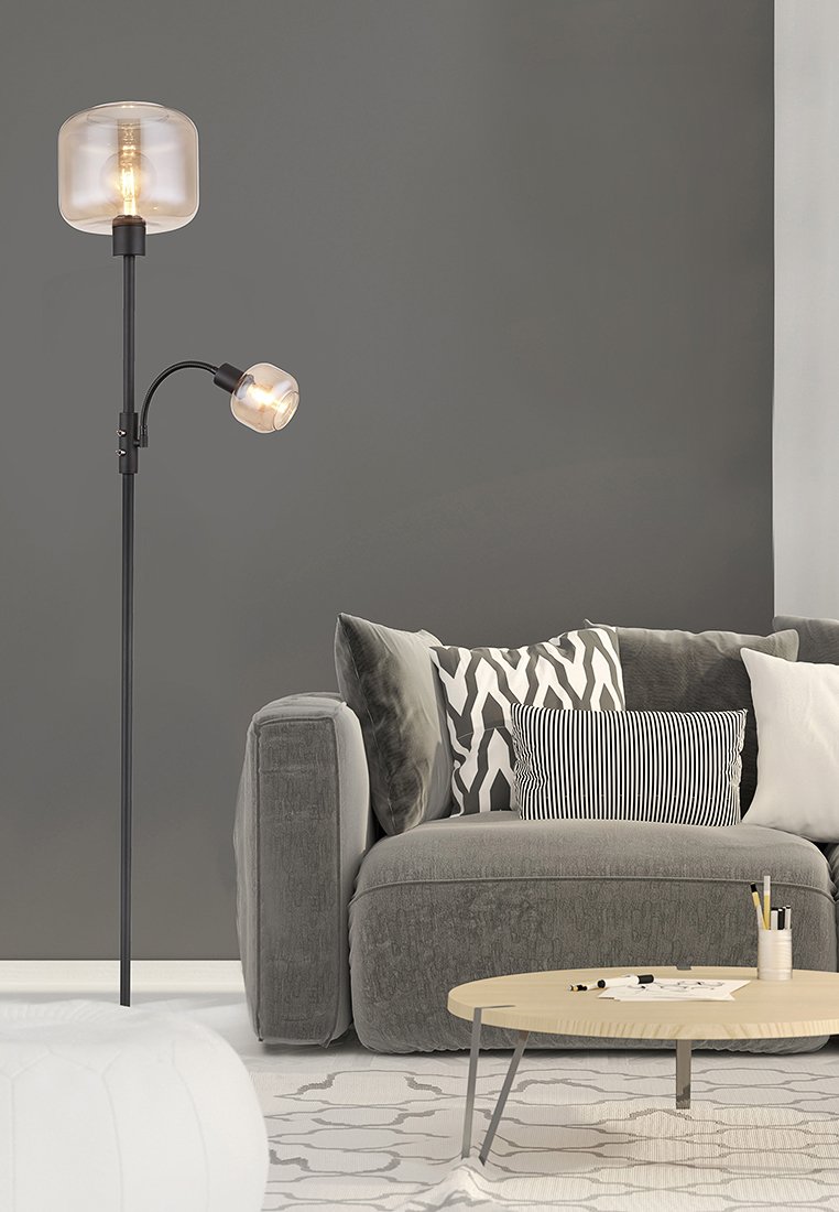 Hohe schwarze Stehlampe mit zwei beleuchteten Glühbirnen neben einem grauen Sofa mit gemusterten Kissen und einem runden Holztisch, auf dem Marker und Papier liegen.