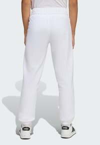 Biele tepláky s elastickým pásom, zúženým strihom a mäkkou textúrou. Nosené s čierno-bielymi teniskami so tromi pruhmi.