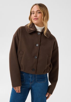 Femme portant une veste marron boutonnée sur un haut blanc et un jean bleu, debout devant un fond clair uni.