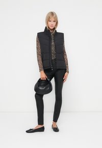 Zwarte gewatteerde vest, top met luipaardprint, zwarte slim-fit broek, zwarte flats en een zwarte handtas. Model staat tegen een witte achtergrond.