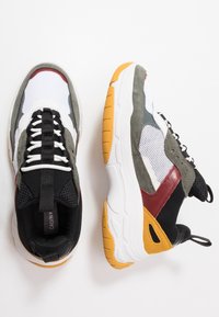 Baskets multicolores présentant un mélange de matériaux : maille blanche, suède gris, textile noir et accents rouges. Semelle épaisse et texturée en blanc et jaune.