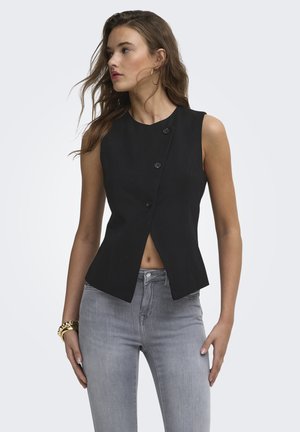 Femme portant un haut noir sans manches boutonné et un jean gris, regardant vers la gauche avec des cheveux lâches et ondulés, un bracelet en or et des boucles d’oreilles créoles.