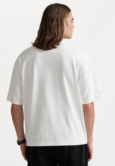 Persona con cabello castaño hasta los hombros, vestida con una camisa blanca de manga corta y suelta, y pantalones negros, vista desde atrás sobre un fondo blanco.