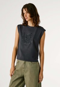 Sivi bezrukavi crop top od pamuka, s grafičkim printom koji uključuje tekst i krila u crnoj boji. Uparen s labavim zelenim hlačama.