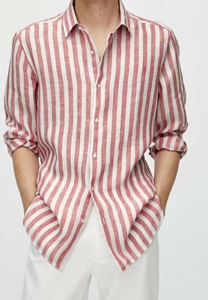 Camicia di lino a righe rosse e bianche con colletto button-down, maniche arrotolate e orlo curvo, indossata sopra pantaloni bianchi.