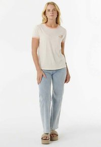 T-shirt beige clair avec bordure corail, présentant un petit motif floral sur le côté gauche de la poitrine. Associé à un jean délavé clair et des sandales compensées couleur camel.