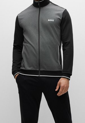 Schwarze und graue Zip-up-Trainingsjacke mit einem hohen Kragen, ausgestattet mit einem weißen Logo und kontrastierenden Paspeln entlang des Reißverschlusses und der Bündchen.