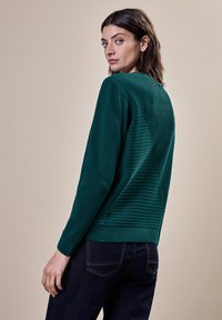 Pull vert à côtes avec un motif texturé, col rond et ourlet ajusté, associé à un jean bleu foncé. Le modèle est vu de dos.