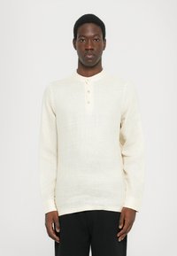 Lys beige langermet skjorte med rund krage og tre knapper. Teksturert stoff med et subtilt mønster. Kombinert med svarte bukser.