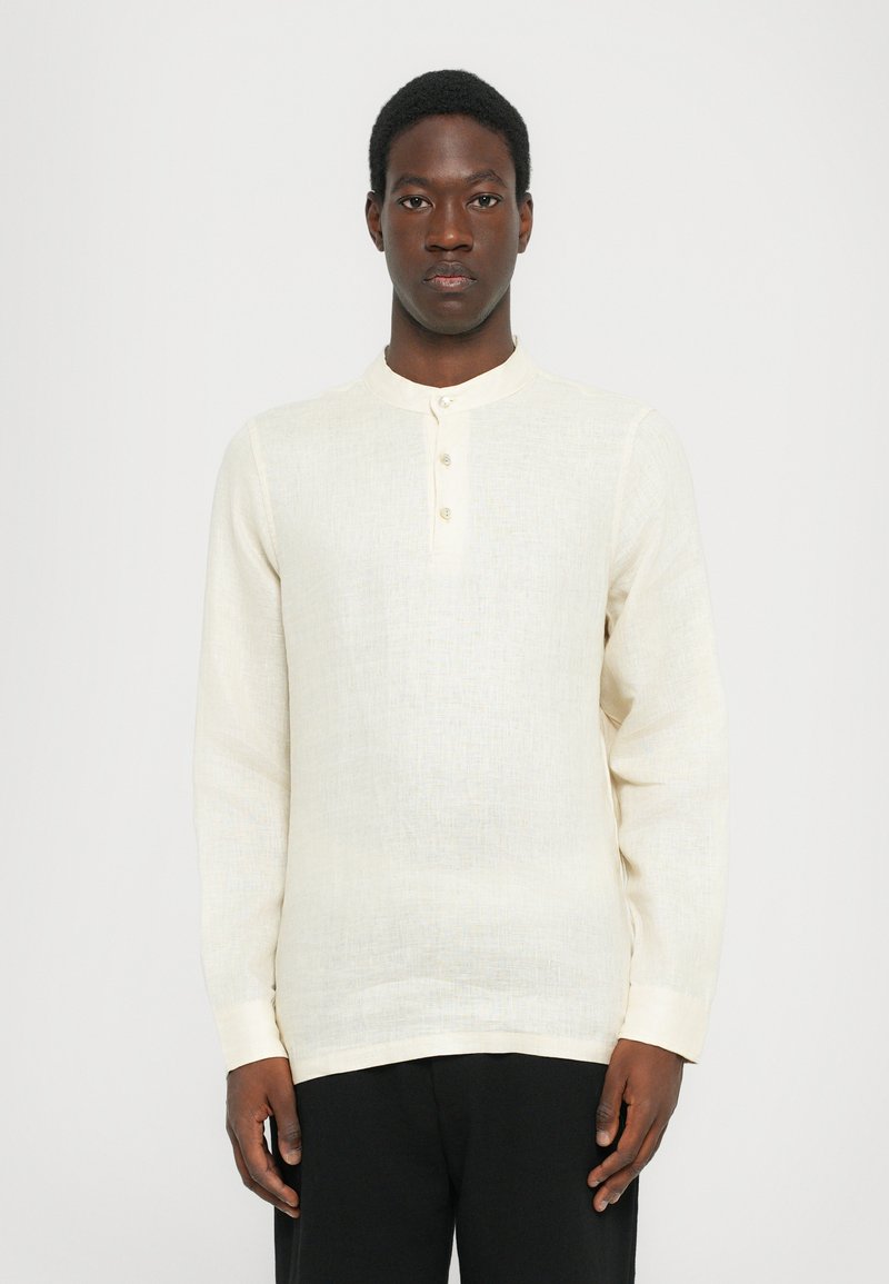 Lys beige langermet skjorte med rund krage og tre knapper. Teksturert stoff med et subtilt mønster. Kombinert med svarte bukser.
