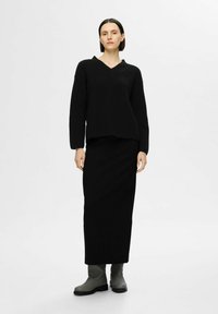 Maglione nero lavorato a maglia con scollatura a V e texture a coste, abbinato a una gonna nera lunga fino al pavimento e stivaletti grigi alla caviglia. Design semplice e minimalista.