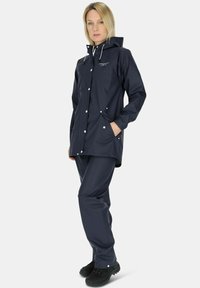 Granatowa wodoodporna kurtka przeciwdeszczowa z kapturem, z zatrzaskami i kieszeniami, zestawiona z pasującymi granatowymi spodniami i czarnym obuwiem.
