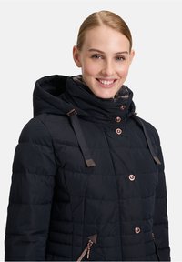 Giacca puffer trapuntata blu navy con cappuccio, caratterizzata da dettagli con bottoni marroni, cinturini regolabili e tasche con zip. Fodera interna morbida visibile.