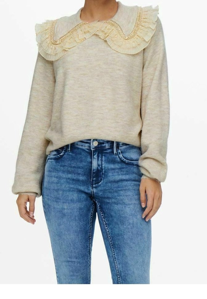 Pull beige avec un grand col en dentelle plissée associé à un jean bleu en denim taille haute sur une personne debout.