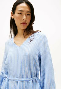 Lyseblå, stribet hørtop med V-udskæring og lange ærmer. Har en bælte i taljen og tekstureret stof med fine, vandrette linjer.