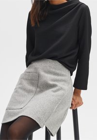 Schwarzes, strukturiertes Langarmoberteil kombiniert mit einem grauen, gestreiften Rock mit großer Tasche und asymmetrischem Saum. Modell sitzt auf einem Hocker.