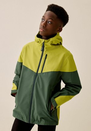 Junge Person trägt eine zweifarbige grüne wasserdichte Jacke mit den Händen in den Taschen und schaut leicht zur Seite vor einem einfarbigen Hintergrund.