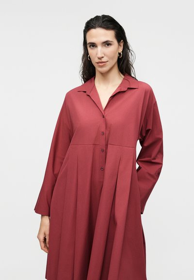 Γυναίκα με σκούρα μαλλιά φοράει χρυσούς κρίκους στα αυτιά και ένα χαλαρό, μακρυμάνικο, σκούρο κόκκινο φόρεμα με κουμπιά, σε ανοιχτό φόντο.