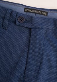 Granatowe, teksturowane spodnie z zapięciem na guzik i pasem. Metka brzmi "DRYKORN FOR BEAUTIFUL PEOPLE." Posiadają subtelne wzory.