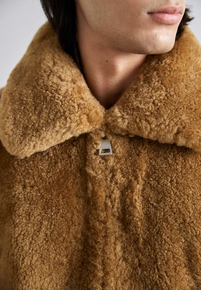 Caban din blană de miel maro, cu guler larg, având un detaliu cu cataramă argintie. Textura este moale și pufoasă, cu un aspect cald și confortabil.