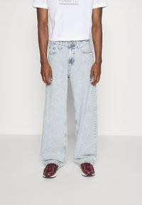 Světlé modré džínsy s širokými nohavicemi z denimu s vybledlým vzhledem. Nošeny s bílým tričkem a tmavě červenými teniskami.